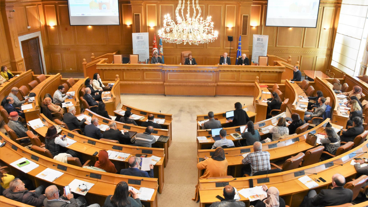 Vue d'ensemble d'une assemblée plénière dans une salle de conférence aux boiseries classiques, montrant des participants assis en arc de cercle lors d'une session de travail sur le dialogue régional..