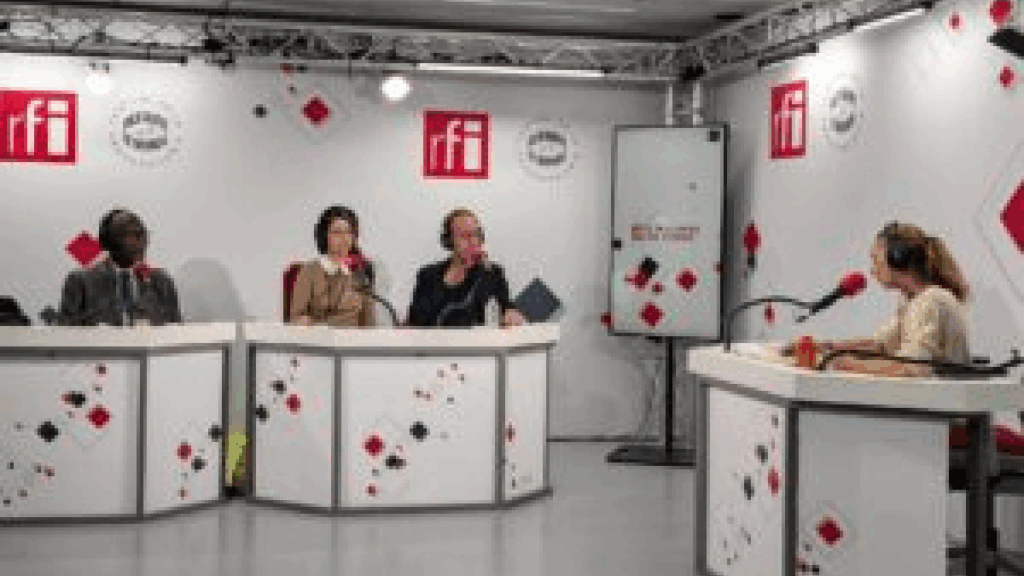 Studio de la radio RFI montrant quatre intervenants assis autour d'une table blanche avec des micros lors de l'enregistrement de l'émission "8 milliards de voisins".