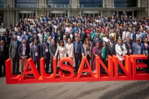 Grand groupe de représentants municipaux rassemblés devant le Centre de Congrès de Lausanne autour d’un grand lettrage rouge “LAUSANNE”.